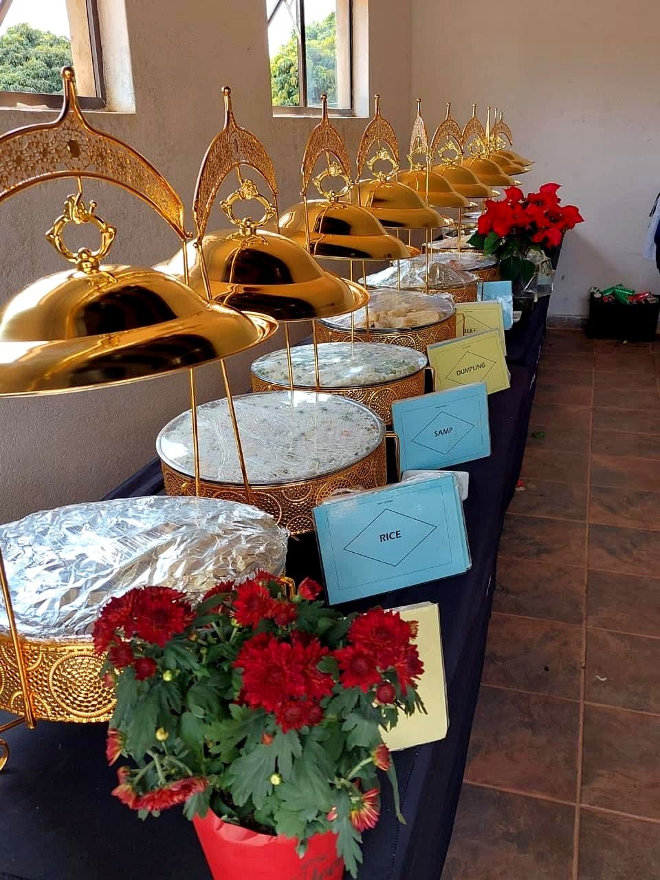 A row of gold serving dishes on a black-clothed buffet table, labelled rice, samp, dumpling and beef, with red flowers